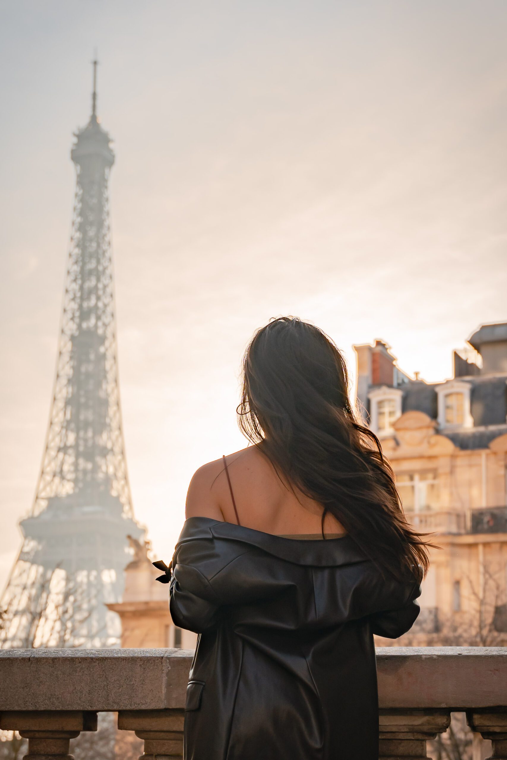 Photographe en Ile-de-France, Femme aux cheveux longs de dos face à la tour Eiffel à Paris avec sa veste qui lui tombe des épaules