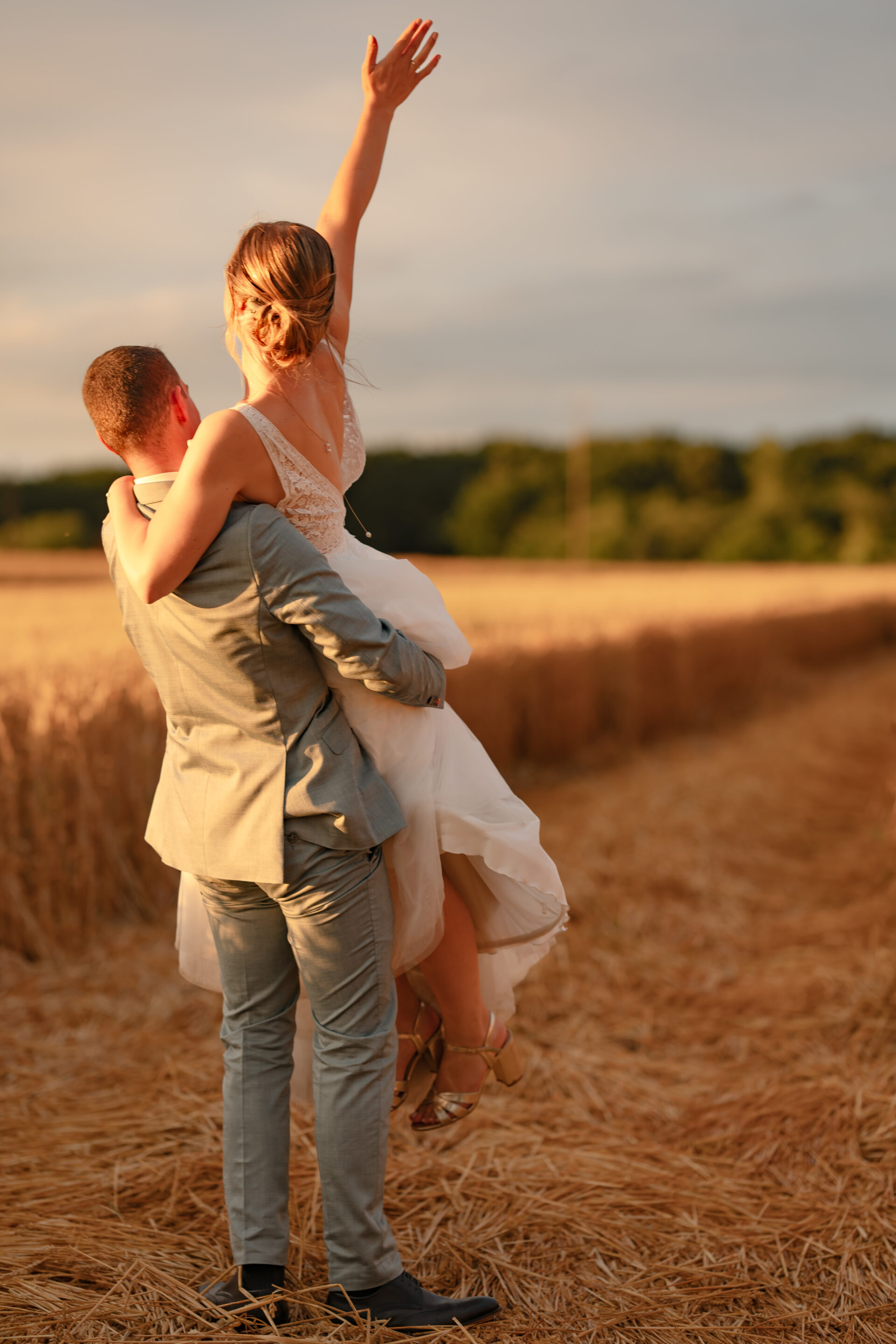 Couple de jeunes mariés dans un champ de blé. Le marié porte sa femme qui lève un bras vers le ciel